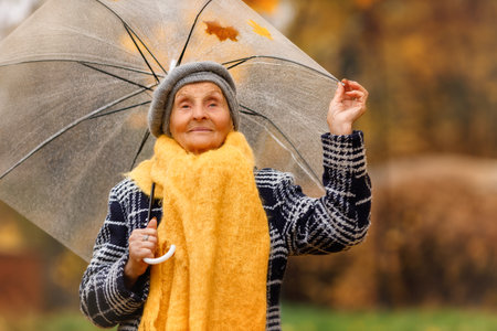 A senior woman over 80, smiling and spinning a transparent umbrella outdoors. She looks at the camera while surrounded by yellow autumn leaves. A heartwarming moment of joy in nature.の写真素材