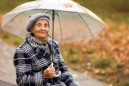 An elderly woman in a beret sits on a bench in autumn, holding a transparent umbrella and looking directly at the camera. Peaceful and warm autumn mood.の写真素材