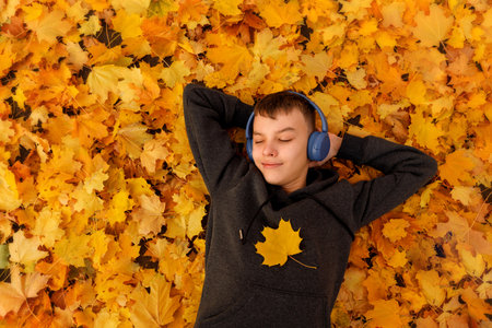 A teenage boy sits on a bench with his arms stretched along the backrest, eyes closed, fully immersed in the music playing in his headphones.の写真素材