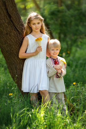 Little girl and boy holding dandelion bouquets, standing together under a tree in a green park. The boy wears traditional embroidered clothing, and the girl wears a white summer dress.の写真素材