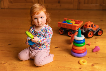Adorable red-haired toddler girl sitting on a wooden floor in a cozy room, surrounded by bright...