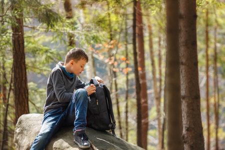 Young boy sitting on a large rock in a forest, opening his backpack. Child dressed in casual outdoor clothing, Concept of hiking, exploration, and childhood adventure in nature.の写真素材