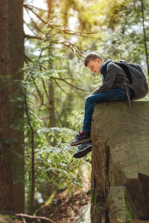 A boy sits on a large rock in the forest during a hike, wearing casual clothes and holding a backpack. Sunlight filters through the trees, creating a peaceful outdoor atmosphere.の写真素材