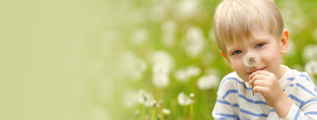 Blonde boy holding a dandelion and smiling in a blooming meadow full of white seed heads on a sunny day.の写真素材
