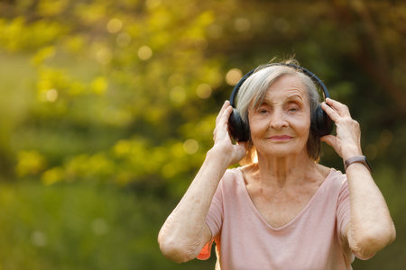 Elderly gray-haired woman stands outdoors with eyes closed, gently holding wireless headphones,...