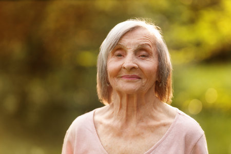 Portrait of a gray-haired elderly woman smiling directly at the camera, standing outdoors in soft...