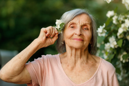 A senior woman gently presses a jasmine twig to her temple, lost in thought, surrounded by blooming...