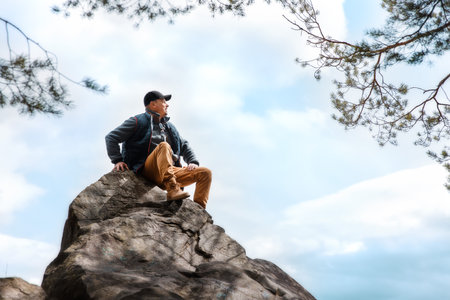 Man in cap and vest sits on a top large rock, looking out at view. Hiker resting during outdoor adventure under cloudy sky.の写真素材