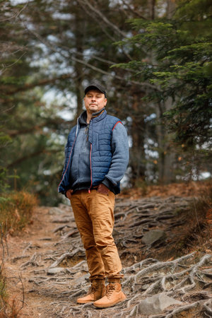 Man in cap and blue vest standing on forest trail with tree roots. Hiking, nature, outdoor activity.