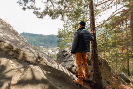 Rear view of a man wearing outdoor clothes and hiking boots standing on a large rock, resting his...