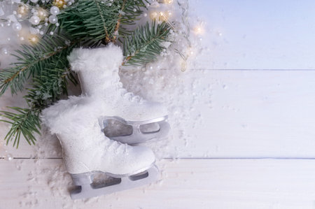 Close-up, high-angle view of miniature white ice skates with fur trim resting on artificial snow next to pine branches and string lights, set against a white wooden background.の写真素材