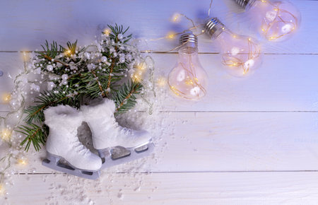 Top-down view of a pair of miniature white glitter ice skates placed on pine branches, surrounded by sparkling string lights and snow on a white wooden plank background.