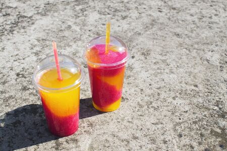 Multi-colored fruit cocktail of crushed frozen ice on a concrete background. Place for text, layout for design. Cooling orange-pink non-alcoholic takeaway sweet drink in a plastic glass.の写真素材