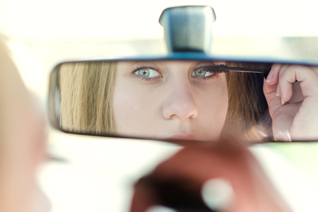 Woman putting make up in a car. Pretty young woman looking in mirror. Dangerous situationの写真素材