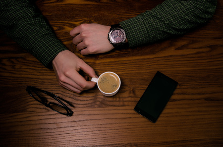 Coffee break. Man hand keep a cup of coffee on a wooden table. Phone and glasses. Business situation.の写真素材