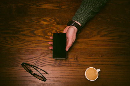 Man hand keep smart phone on a wooden table. Phone and glasses. Business situation. Coffee breakの写真素材