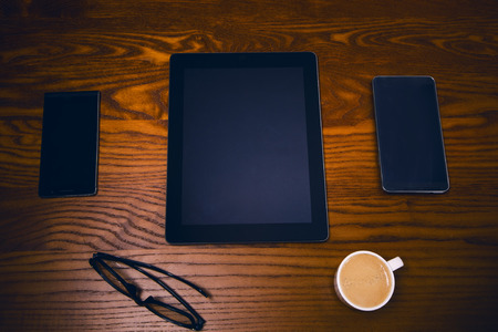 tablet and two mobiles on a wooden table. Cup of coffee and glasses. Concept of technologyの写真素材