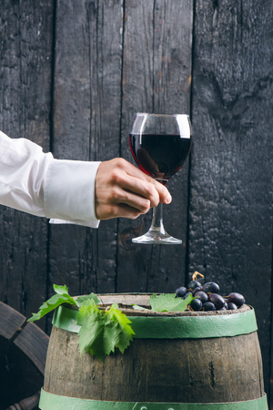 Man hand holds a glass of red wine. Wine on a wooden barrel. Burnt, black wooden background. Vintage. Copyspace for a text. Grapes and green vine.の写真素材