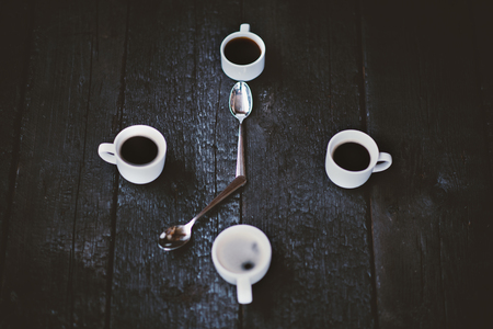 Coffee cups aranged as a clock face on a wooden black background. Four espresso mugs like a symbol of time with two spoons. Time for coffee. Breakfast time. Good morning concept.の写真素材