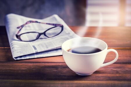 Coffee cup with newspaper and glasses in background on wooden table. Vintage tone photo with sunlight filter effect.の写真素材