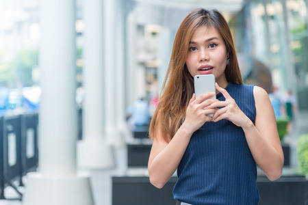 Beautiful young Asian woman feeling surprised while holding the phone outdoorの写真素材