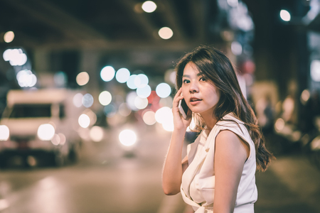 Beautiful Asian woman talk to smartphone at night with blurred city traffic in backgroundの写真素材