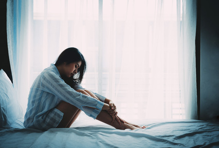 Unhappy beautiful Asian woman sitting on a bed looking sad and lonelyの写真素材