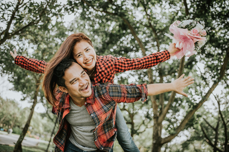Laughing Asian man giving piggyback to his girlfriend with flower bouquet in hand at the park. Arms stretched out together. Happy couple life conceptの写真素材