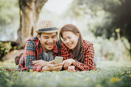 Happy Asian couple lying on green grass laugh and smile while using smartphone.の写真素材