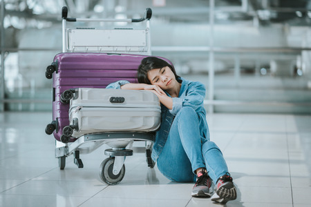 Young Asian woman passenger sleeping with luggage trolley at an airport terminal waitng for departure flightの写真素材