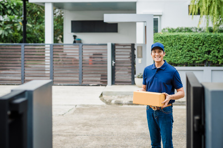 Happy Asian deliveryman holding cardboard box in front of house. Working with service mind.の写真素材