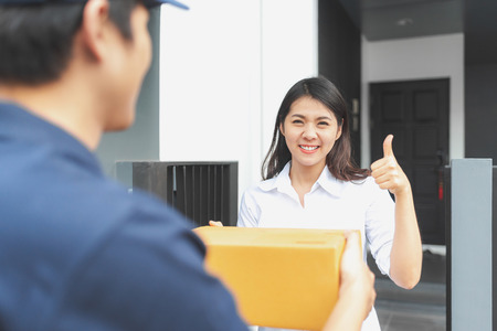 Happy Asian woman giving thumb up after received cardboard package from delivery man in front of her homeの写真素材