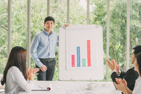 Confident young man standing next to flipchart and smiling while his colleagues giving applaudingの写真素材