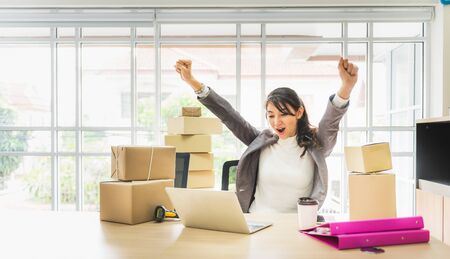 Happy successful business woman with arms up working with laptop and cardboard box on table in home office. business online and delivery concept.の写真素材