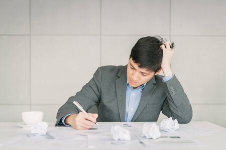 Young Asian businessman looking exhausted and tried from overwork with crumpled papers on his deskの写真素材