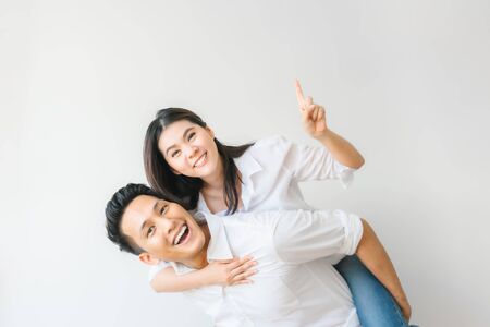 Happy Asian couple in love having fun piggyback ride and point at empty copyspace on white wall backgroundの写真素材
