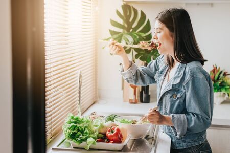 Happy Asian woman eating fresh vegetables while preparing salad in kitchen. Healthy food conceptの写真素材
