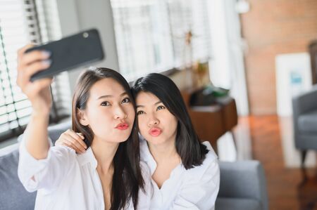Two happy beautiful Asian girl friends using smartphone selfie in living room at home with smiling...