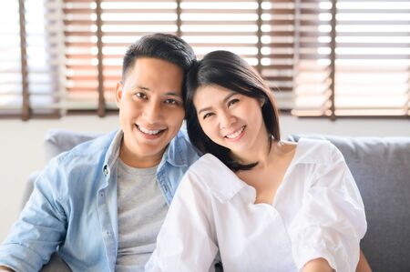 Portrait of happy Asian couple in love smiling relaxing on sofa at homeの写真素材