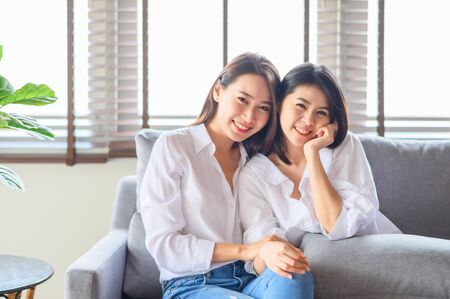 Two cute young happy Asian women smiling and laughing while sitting on sofa in living roomの写真素材