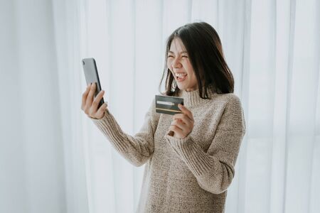 Happy and excited Asian woman using credit card for online shopping with smartphone in her house