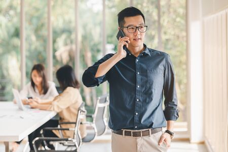 Portrait of smiling Asian businessman using smartphone to discuss business plan in meeting room at the office with colleague in backgroundの写真素材