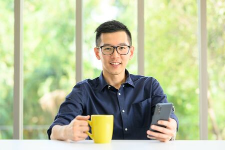 Happy smiling Asian man using smartphone mobile in modern office looking at camera
