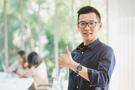 Happy Asian businessman smiling with thumbs up gesture with colleague in background in meeting room