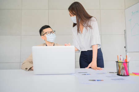 Asian man and woman entrepreneur wears face mask discuss business project in laptop in meeting room...