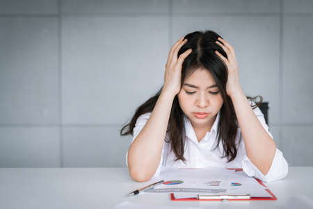Asian woman looking stressed and tried from work with her hand on her head at working tableの写真素材