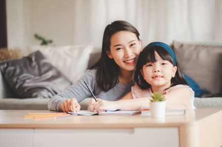 Happy Asian family mother and daughter sitting on sofa reading a book in living room at home