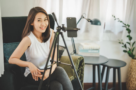 Happy healthy smiling young Asian woman blogger looking at camera while using smartphone for recording live video vlog at homeの写真素材