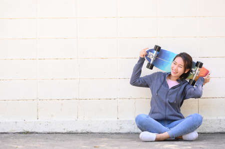 Happy cheerful Asian woman in casual outfit with surfskate sitting against concrete wall. active...