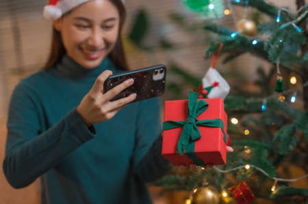 Happy Asian woman using smartphone mobile taking selfie with Christmas gift box with Christmas tree in backgroundの写真素材